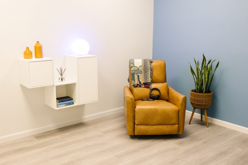 A cozy therapy room with a mustard-yellow recliner, a patterned blanket draped over the back, black headphones on the seat, and a tall potted snake plant beside it. Floating white shelves with decorative vases, books, and a glowing orb lamp are mounted on the wall.
