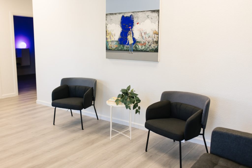 A modern waiting area with two black chairs on either side of a small white table holding books and a plant. A colorful painting of a large blue cat with a girl holding a balloon is hung on the wall above. In the background, a softly lit room glows purple-blue.