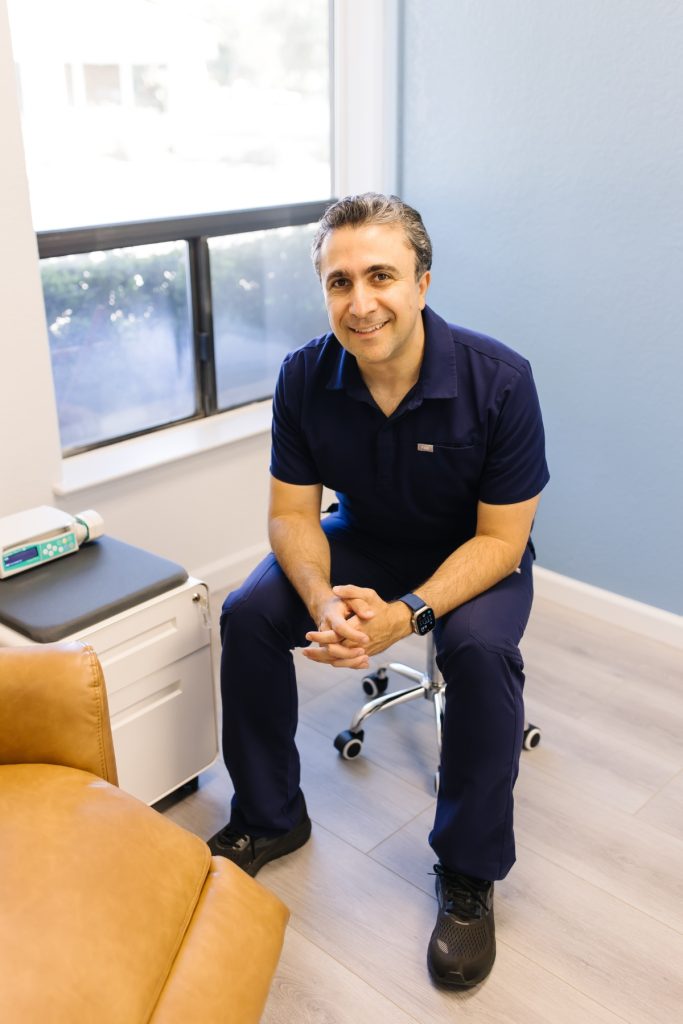 A smiling healthcare professional in navy scrubs sits on a stool next to a tan recliner in a bright clinic room with light wood floors and a window. Medical equipment rests on a small cabinet nearby.