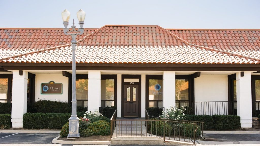 The exterior of Bluvera Clinic, a single-story building with a terracotta tile roof, white walls, and black-trimmed windows. A ramp leads to the glass front door with the suite number 103, flanked by neatly trimmed bushes and flowers.
