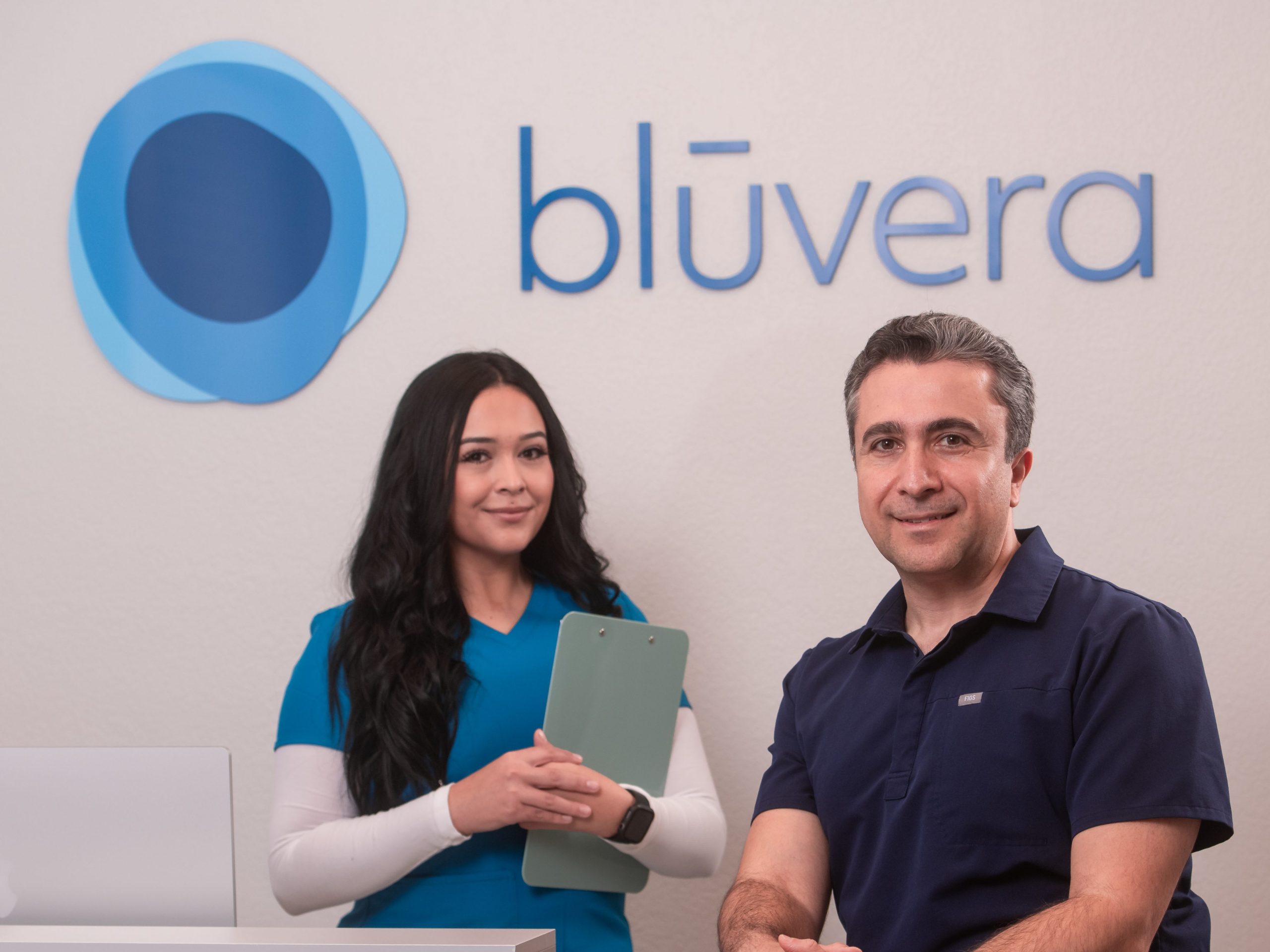 Two healthcare professionals pose at a reception desk beneath a blūvera logo - one seated in navy scrubs, the other standing in teal scrubs holding a clipboard, with a laptop on the counter.